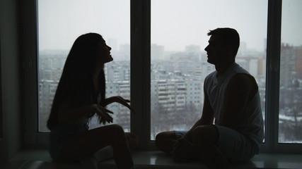 Young family couple sitting on the windowsill, emotionally talking, arguing and gesticulating