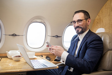 Portrait of contented diplomat sitting at tray table in airplane and holding cellphone in hands, he...
