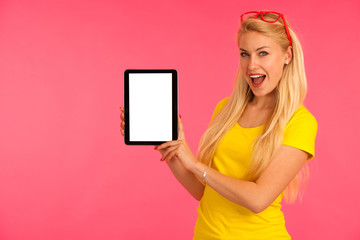 Beautiful young woman sin yellow t shirt holds tablet and surfs internet over pink background