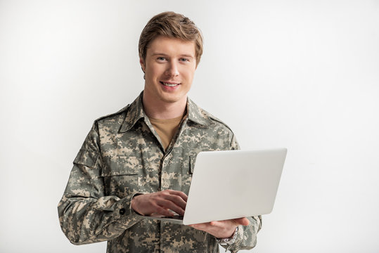 Glad Military Young Man Looking At Camera With Calm Smile. He Is Holding Laptop In Hand And Typing. Isolated On Background