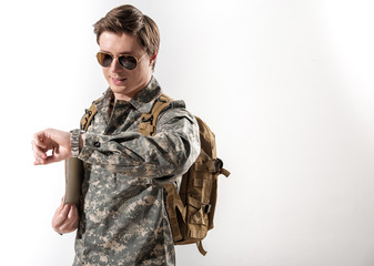 Confident military guy looking at his watch and smiling. He is having folder at hand and backpack on his shoulders. Isolated on background. Copy space in right side