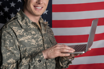 Joyful young military man smiling and having device in his hands while typing. Flag on background