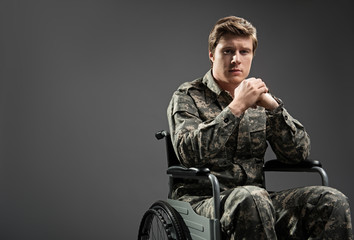 Upset young male looking at camera with sad look. He is sitting in invalid chairing wearing...