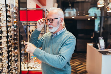 Handsome senior man choosing eyeglasses frame in optical store. 