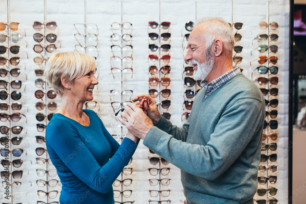 Wall mural Happy senior couple choosing together eyeglasses frame in optical store. 