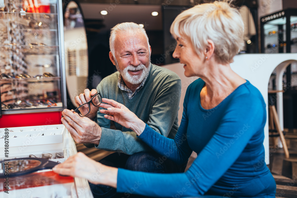 Wall mural Happy senior couple choosing together eyeglasses frame in optical store. 