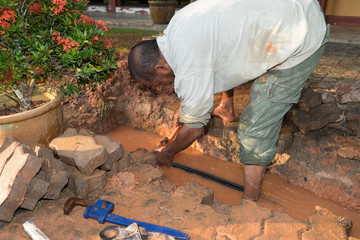 Plumber using a pipe wrench to repair a leaking pipe under the interlocking bricks walkway.