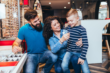 Happy mother and father choosing eyeglasses frame for their son in optical store. 