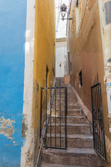 A very narrow walkway with stone steps, going up-hill, with a black iron gate at the entrance, and colorful walls of blue and yellow, in Guanajuato, Mexico - 191813003