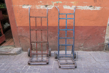 Isolated shot of two metal dollies, one red and one blue, against a red and orange exterior wall, with stone pavement, in Mexico - 191813012