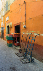 Market with bright orange with red border exterior wall, with a red and blue hand truck leaning against the wall, some crates filled with fruit and stone pavement, in Guanajuato, Mexico