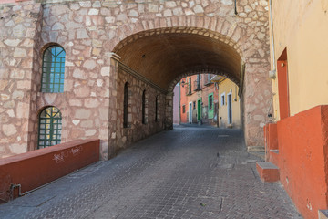 A stone paved street going up a hill and through a stone tunnel with two arched windows on the stone wall, next to the tunnel entrance, and colorful houses, in Guanajuato, Mexico