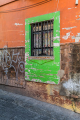 A funky green frame around a double window, with iron bars, a stone wall with orange paint, some graffiti and a stone street, in Guanajuato, Mexico - 191813038