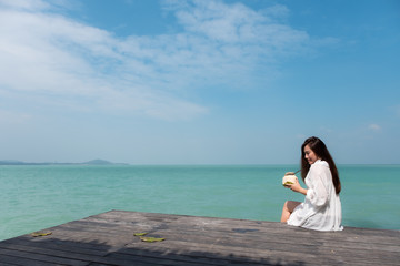 A beautiful asian woman on white dress sitting at the terrace drinking coconut juice with sea and blue sky background with feeling relaxed