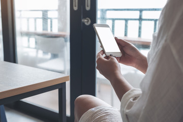 Mockup image of a woman's hands holding white mobile phone with blank black desktop screen in loft cafe