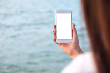 Mockup image of a woman's hand holding white mobile phone with blank desktop screen by the sea and blue sky background