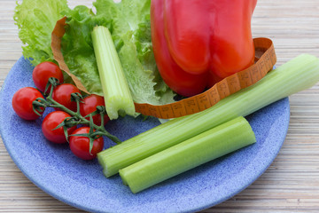 big celery,tape measure for diet, fresh green lettuce leaves next to red pepper and red cherry tomatoes lying on a blue plate, wooden table background
