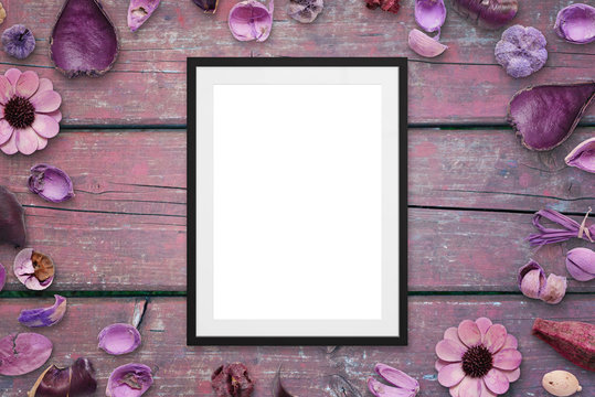 Picture frame on pink wooden desk surrounded with flower decorations. Top view.