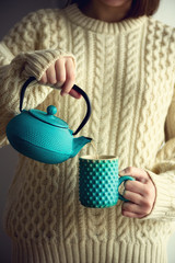 Woman in warm knitted woolen sweater holds turquoise teapot and pouring herbal tea into handmade cup. Copy space. Winter and Christmas holidays concept.