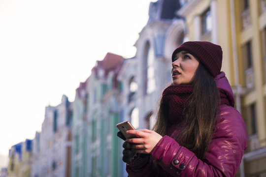 Pretty Brunette Girl Wearing Purple Winter Coat, Hat and Scarf, Walking by European Street at Winter, make Photos on Her Smartphone and Making Selfie