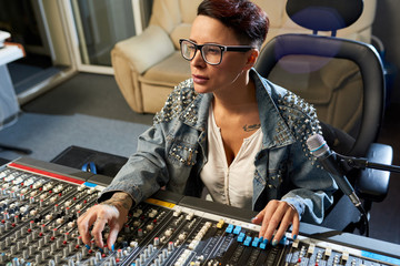 Thoughtful modern female audio engineer concentrated on mixing sounds while moving faders and twisting SSL channels on digital audio channel mixer in studio