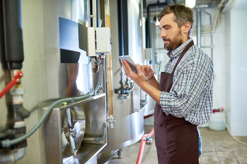 Serious thoughtful handsome brewing engineer in apron examining equipment using modern device while working at factory