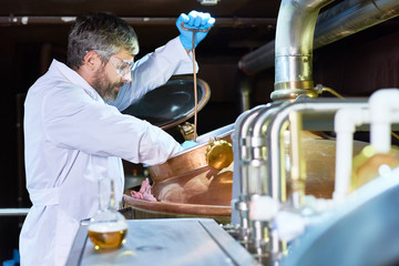 Serious concentrated handsome middle-aged brewer wearing protective eyewear and gloves examining fermentation in tank while working at craft brewery