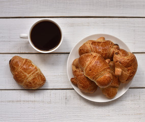 coffee mood. country breakfast. close up of a cup of black coffee and a plate with freshly baked croissants with golden crust on a white retro  wooden table