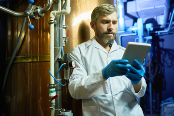 Thoughtful concentrated brewing technician in lab coat using digital tablet while learning specialties of brewery machinery at plant