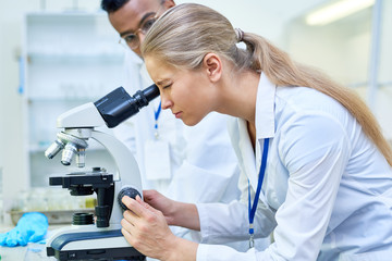 Side view portrait of young female scientist looking in microscope while working on medical research in science laboratory