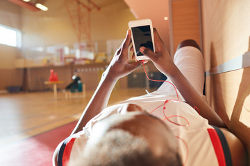 Fototapeta premium Tired African basketball player waiting for training and relaxing on bench enjoying music in earphones and using gadget while checking social media on training court
