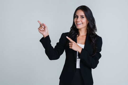 Look here! Beautiful young businesswoman pointing away and looking at camera with smile while standing against white background