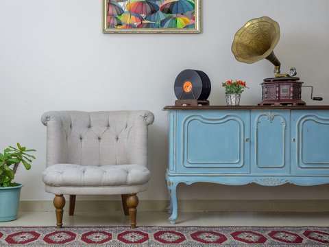 Retro off white armchair, vintage wooden light blue sideboard, old phonograph (gramophone) and vinyl records on background of beige wall, tiled porcelain floor, and red carpet