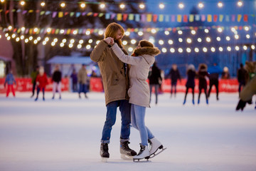 Theme ice skating rink and loving couple. meeting young, stylish people ride by hand in crowd on city skating rink lit by light bulbs and lights. Ice skating in winter for Christmas on ice arena - obrazy, fototapety, plakaty