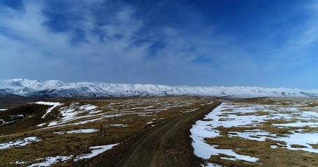 Aerial photograph of the Tibetan Plateau showing grassland partially covered by snow, a dirt road extending towards the horizon and a snow-covered mountain range in the background.
