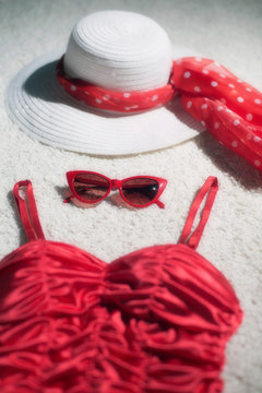 Vintage summer hat, red sunglasses and swimsuit displayed on white carpet floor.