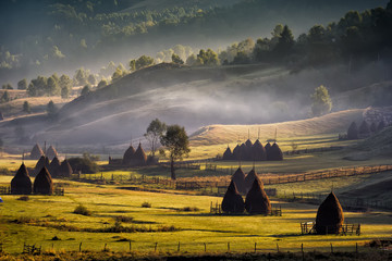 Beautiful rural mountain landscape in the morning light with fog, old houses and haystacks,...