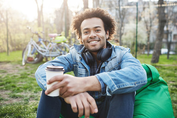 Naklejka na ściany i meble Happy and positive young male student with afro-hairstyle in trendy clothes sitting in park while smiling broadly and drinking coffee. Boyfriend suggest to have a rest after spending day riding bike