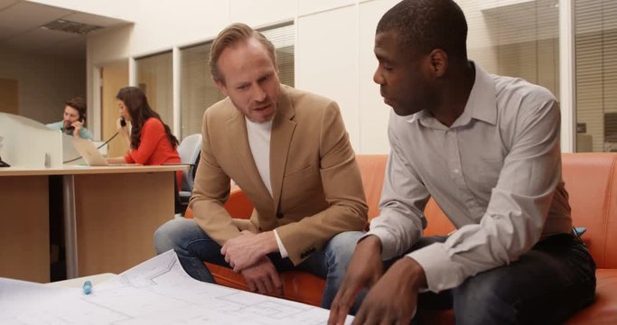 4K Businessmen discussing floor plans for new building development in the office, with staff working in the background. Slow motion.