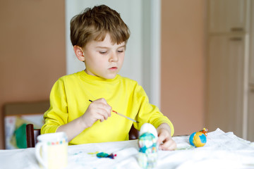 Little blond kid boy coloring eggs for Easter holiday in domestic kitchen, indoors.