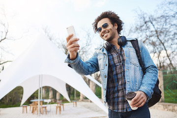 Outdoor portarit of happy sincere african american man in stylish glasses and denim coat, holding cup of coffee and taking photo on smartphone while smiling broadly during walk in park.