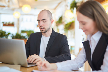Obraz premium Young businessman sitting in cafe in front of laptop and working online with colleague near by