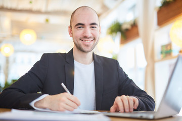 Young smiling businessman making notes while sitting in front of laptop in cafe and looking at camera