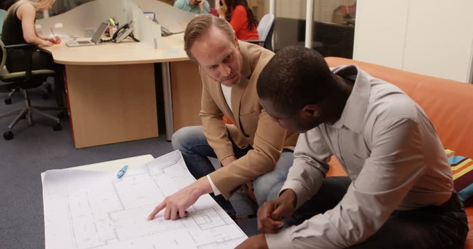 4K Businessmen discussing floor plans for new building development in the office, with staff working in the background. Slow motion.