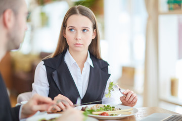 Serious young businesswoman sitting by table in restaurant by lunch and listening to her colleague explanation