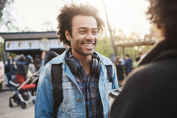 Emotive charming dark-skinned guy with afro haircut and sincere smile talking to his girlfriend, deciding where to go next while walking in park or street, wearing trendy outfit.