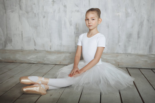 A little adorable young ballerina in white dress and point shoes sitting on woody floor and gray studio background posing on camera