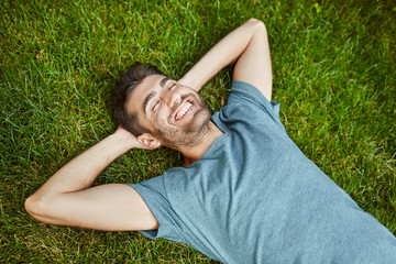 Positive emotions. Young beautiful bearded caucasian male in blue t-shirt lying on grass smiling with teeth, laughing, relaxing outside in summer morning with happy face expression.