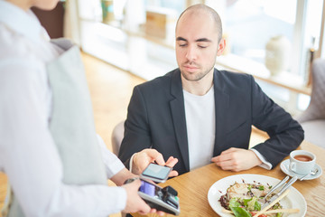 Modern businessman holding his smartphone over payment terminal in cafe