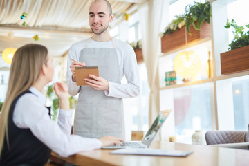 Smiling waiter writing down order of client in notepad during working day